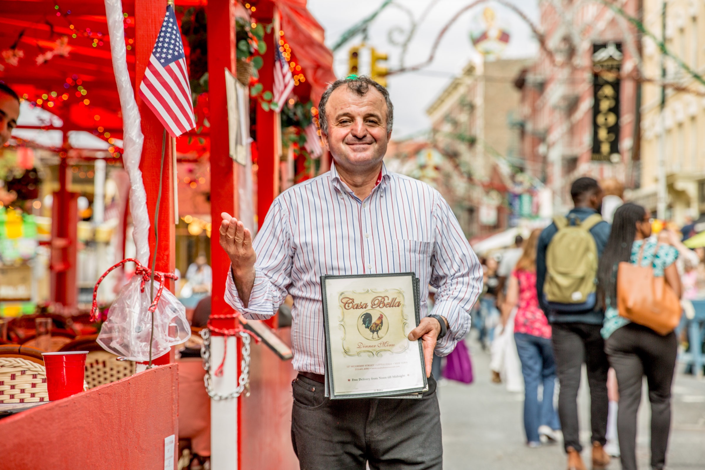 A host standing outside a restaurant holding a restaurant menu during the Feast of San Genaro in NYC.